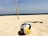 A volleyball rests on a sandy beach with clear skies above. Bamboo poles with a net are set up in the background, suggesting a beach volleyball area. Distant boats and people can be seen near the shoreline.