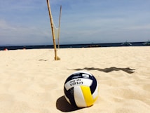 A volleyball rests on a sandy beach with clear skies above. Bamboo poles with a net are set up in the background, suggesting a beach volleyball area. Distant boats and people can be seen near the shoreline.