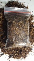 A clear plastic bag filled with dried, shredded brown material, possibly tobacco or a similar substance, rests on a surface. Loose pieces of the material are scattered around the bag.