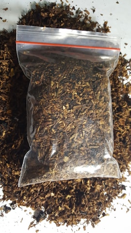 A clear plastic bag filled with dried, shredded brown material, possibly tobacco or a similar substance, rests on a surface. Loose pieces of the material are scattered around the bag.