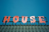 A brightly colored sign spelling out the word 'HOUSE' in red letters with a jagged, playful font, each letter adorned with small round yellow lights. The sign is mounted on a metallic structure above a roof with green tiles, set against a clear blue sky.