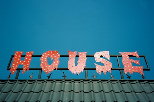 A brightly colored sign spelling out the word 'HOUSE' in red letters with a jagged, playful font, each letter adorned with small round yellow lights. The sign is mounted on a metallic structure above a roof with green tiles, set against a clear blue sky.