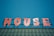 A brightly colored sign spelling out the word 'HOUSE' in red letters with a jagged, playful font, each letter adorned with small round yellow lights. The sign is mounted on a metallic structure above a roof with green tiles, set against a clear blue sky.