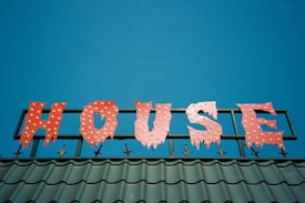 A brightly colored sign spelling out the word 'HOUSE' in red letters with a jagged, playful font, each letter adorned with small round yellow lights. The sign is mounted on a metallic structure above a roof with green tiles, set against a clear blue sky.