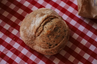 A freshly baked round loaf of rustic bread rests on a red and white checkered tablecloth. Nearby, part of a brown paper bag is visible, partially folded.