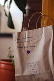 A brown paper bag with handwritten text and a purple heart on it. The text is in Portuguese, expressing well wishes for the weekend. The bag is labeled with 'CAIPORA & DALILA' in blue ink. There's an orange pot holding a green plant in the background.
