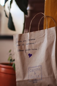 A brown paper bag with handwritten text and a purple heart on it. The text is in Portuguese, expressing well wishes for the weekend. The bag is labeled with 'CAIPORA & DALILA' in blue ink. There's an orange pot holding a green plant in the background.