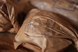 Crinkled brown paper packages are neatly arranged on a wooden surface. The packages have handwritten text on them, possibly denoting a brand, with a clear, simple stamp-like design.