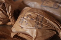 Crinkled brown paper packages are neatly arranged on a wooden surface. The packages have handwritten text on them, possibly denoting a brand, with a clear, simple stamp-like design.