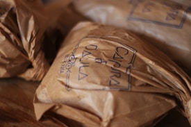 Crinkled brown paper packages are neatly arranged on a wooden surface. The packages have handwritten text on them, possibly denoting a brand, with a clear, simple stamp-like design.