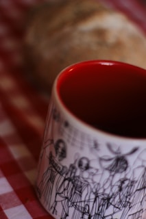 A close-up view of a white mug with artistic black line drawings of people on it. The interior of the mug is painted bright red. In the blurred background, there is a loaf of bread on a red and white checkered cloth.