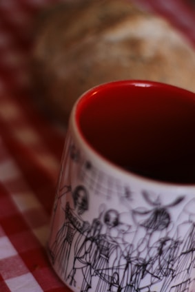 A close-up view of a white mug with artistic black line drawings of people on it. The interior of the mug is painted bright red. In the blurred background, there is a loaf of bread on a red and white checkered cloth.