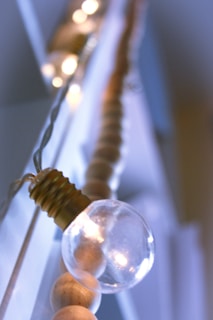 A close-up of a string of lights featuring a prominent clear light bulb with a visible filament, set against a blurred background. The light string includes wooden beads, adding a rustic touch. Soft, ambient lighting creates a warm and cozy atmosphere.