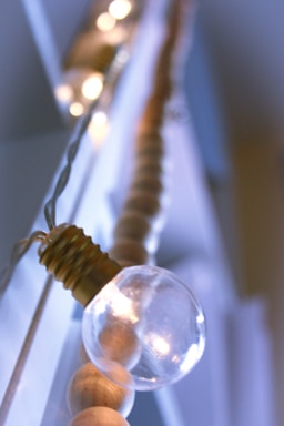 A close-up of a string of lights featuring a prominent clear light bulb with a visible filament, set against a blurred background. The light string includes wooden beads, adding a rustic touch. Soft, ambient lighting creates a warm and cozy atmosphere.