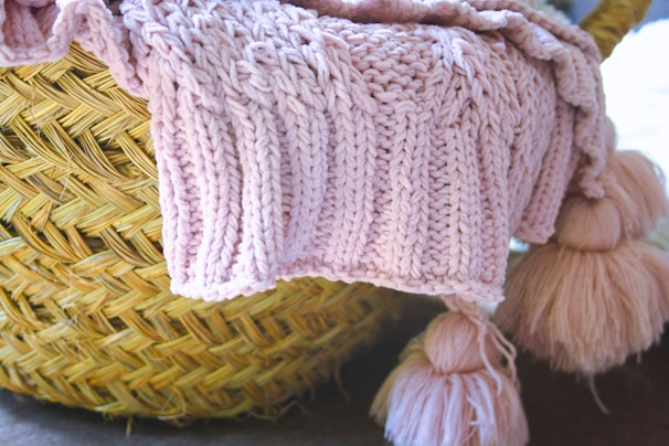 A close-up view of a woven wicker basket with a soft, pink knitted blanket draped over its edge. Attached to the blanket are large, fluffy tassels.