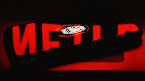 A smartphone reflects the word 'NETFLIX' in bold red letters. In the middle of this reflection, a stylized mask with a distinct expression is visible.