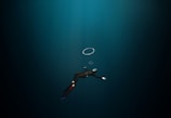 A diver in a dark underwater environment, wearing a wetsuit, fins, and a mask with a snorkel. The diver is swimming downward, and a few air bubbles are visible rising towards the surface.
