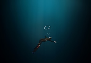 A diver in a dark underwater environment, wearing a wetsuit, fins, and a mask with a snorkel. The diver is swimming downward, and a few air bubbles are visible rising towards the surface.