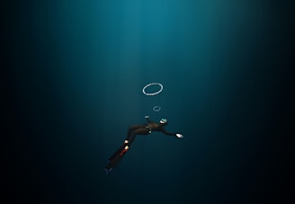 A diver in a dark underwater environment, wearing a wetsuit, fins, and a mask with a snorkel. The diver is swimming downward, and a few air bubbles are visible rising towards the surface.