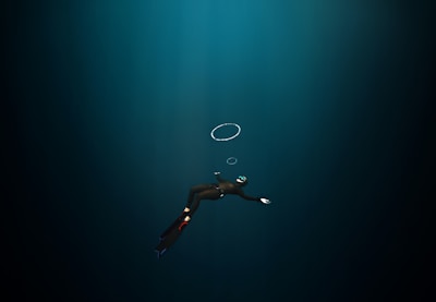 A diver in a dark underwater environment, wearing a wetsuit, fins, and a mask with a snorkel. The diver is swimming downward, and a few air bubbles are visible rising towards the surface.