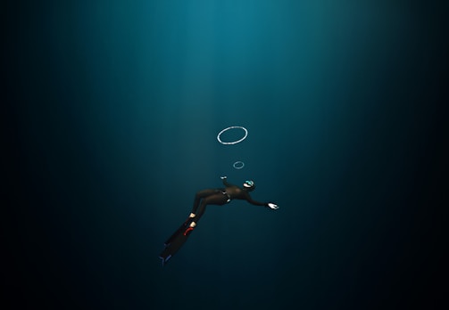 A diver in a dark underwater environment, wearing a wetsuit, fins, and a mask with a snorkel. The diver is swimming downward, and a few air bubbles are visible rising towards the surface.
