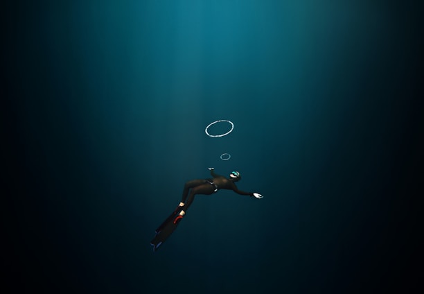 A diver in a dark underwater environment, wearing a wetsuit, fins, and a mask with a snorkel. The diver is swimming downward, and a few air bubbles are visible rising towards the surface.