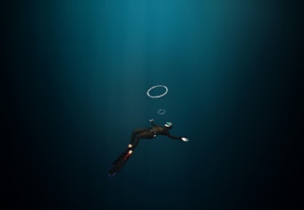 A diver in a dark underwater environment, wearing a wetsuit, fins, and a mask with a snorkel. The diver is swimming downward, and a few air bubbles are visible rising towards the surface.