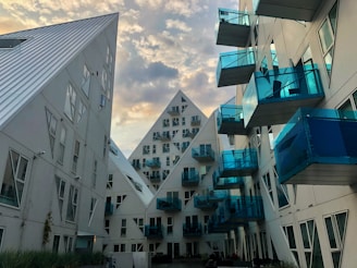 An architectural view featuring modern residential buildings with a striking design. The structures have a geometric, pyramid-like shape and are characterized by white surfaces and numerous blue-tinted glass balconies. The scene is set against a vibrant sky with scattered clouds, enhancing the futuristic look of the buildings.