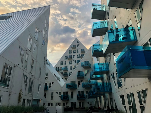 An architectural view featuring modern residential buildings with a striking design. The structures have a geometric, pyramid-like shape and are characterized by white surfaces and numerous blue-tinted glass balconies. The scene is set against a vibrant sky with scattered clouds, enhancing the futuristic look of the buildings.