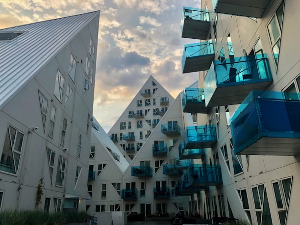 An architectural view featuring modern residential buildings with a striking design. The structures have a geometric, pyramid-like shape and are characterized by white surfaces and numerous blue-tinted glass balconies. The scene is set against a vibrant sky with scattered clouds, enhancing the futuristic look of the buildings.