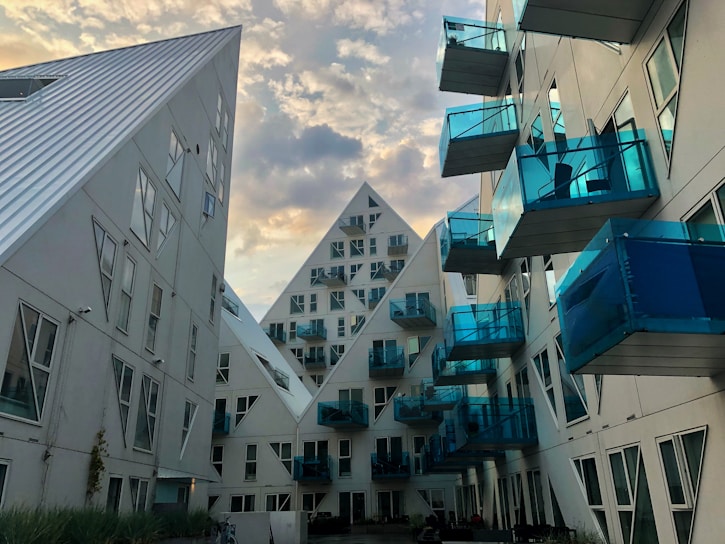 An architectural view featuring modern residential buildings with a striking design. The structures have a geometric, pyramid-like shape and are characterized by white surfaces and numerous blue-tinted glass balconies. The scene is set against a vibrant sky with scattered clouds, enhancing the futuristic look of the buildings.