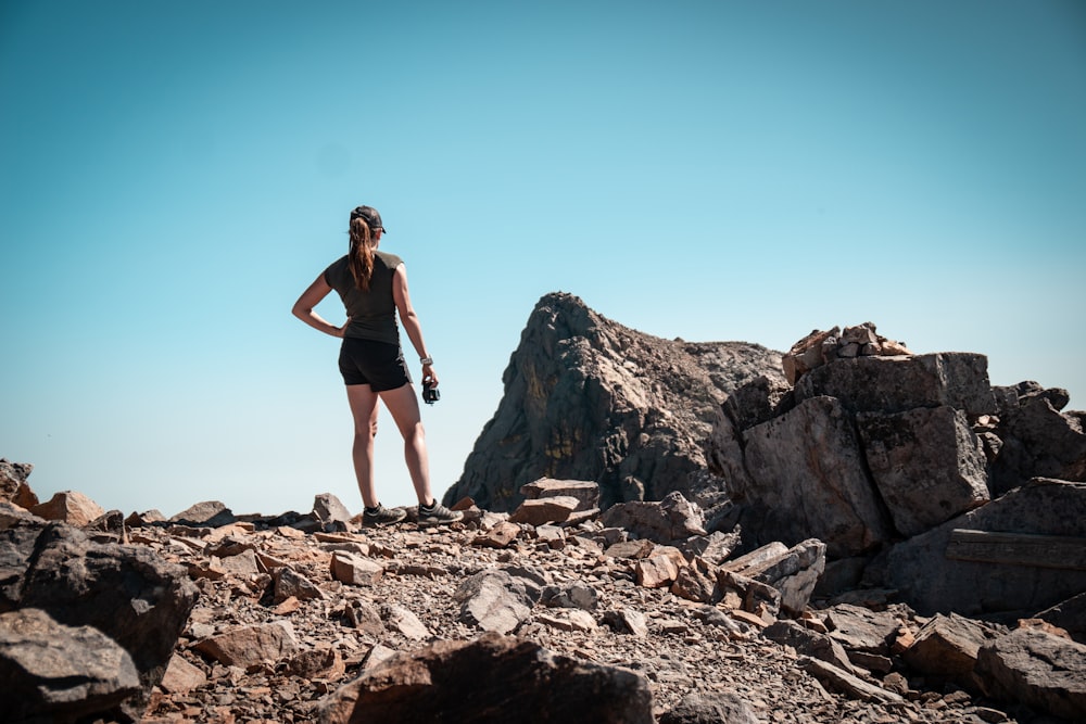 woman in black tank top and black shorts standing on rocky hill under blue sky during