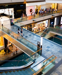 A modern shopping mall interior featuring multiple levels connected by escalators and a glass railing. Shoppers walk along the walkways in front of various high-end retail stores, including Chanel and Guess. The space is lit with bright, ambient lighting, and the floors are made of polished stone tiles.