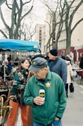 A bustling outdoor market scene with several people interacting and browsing the stalls. One man in a green jacket with a badge is holding a can and looking down. Others are observing items for sale under tents. Background features bare-branched trees and urban buildings.
