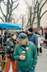 A bustling outdoor market scene with several people interacting and browsing the stalls. One man in a green jacket with a badge is holding a can and looking down. Others are observing items for sale under tents. Background features bare-branched trees and urban buildings.