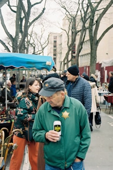 A bustling outdoor market scene with several people interacting and browsing the stalls. One man in a green jacket with a badge is holding a can and looking down. Others are observing items for sale under tents. Background features bare-branched trees and urban buildings.