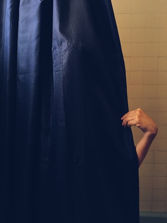 A hand emerges from the side of a dark shower curtain, gripping it gently. The curtain is textured and primarily navy blue, set against a tiled bathroom wall.
