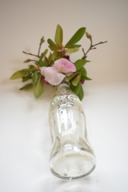 A clear glass Coca-Cola bottle is lying on its side, surrounded by sprigs of greenery and pink flowers. The bottle has a simple, classic design with the Coca-Cola logo and a capacity of 200 ml indicated. The background is a soft, neutral color that complements the natural elements and highlights the transparency of the glass.