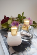 Two elegant bottles with gold caps are placed on a glossy surface, surrounded by a soft focus backdrop of delicate flowers and green leaves. The scene is set in a softly lit environment, enhancing the luxurious appeal of the composition.