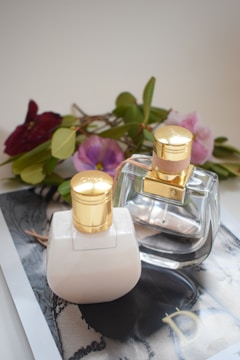 Two elegant bottles with gold caps are placed on a glossy surface, surrounded by a soft focus backdrop of delicate flowers and green leaves. The scene is set in a softly lit environment, enhancing the luxurious appeal of the composition.