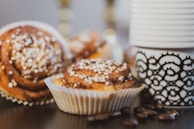 Freshly baked cinnamon rolls topped with pearl sugar are placed next to a black and white patterned coffee cup. Coffee beans are scattered around the wooden surface, adding to the cozy and inviting arrangement.