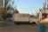 A small white delivery truck is parked on the side of the road. The truck has blue writing on its side, and the surrounding area includes leafless trees and a small red building. The scene is set in an urban environment with a clear sky and afternoon lighting.