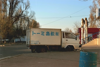 A small white delivery truck is parked on the side of the road. The truck has blue writing on its side, and the surrounding area includes leafless trees and a small red building. The scene is set in an urban environment with a clear sky and afternoon lighting.