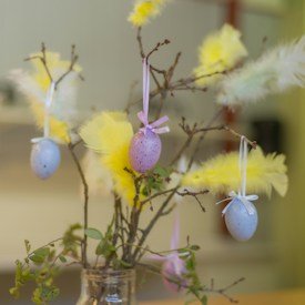 A decorative arrangement featuring branches adorned with pastel-colored speckled eggs tied with ribbons and bright yellow feathers. The branches are placed in a simple glass vase, creating a festive and natural centerpiece.