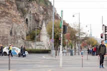 An urban street scene features a rocky cliffside with architectural structures embedded into it. People are walking across a crosswalk, and a police car is present with an officer managing traffic. Trees with autumn foliage line the street, and streetlights and traffic lights are visible.