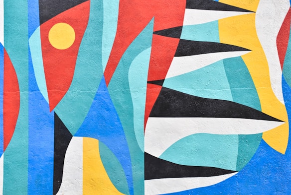 An abstract mural composed of various interlocking geometric shapes. The shapes include triangles, circles, and curved forms in a vivid palette of colors. The arrangement of colors and forms creates a sense of movement and energy.