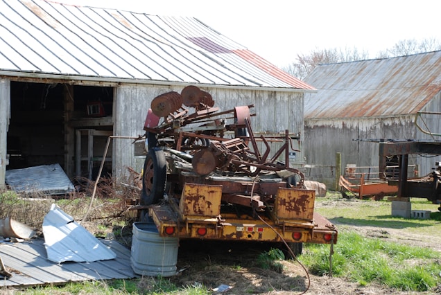 A rusty, old piece of farm machinery is mounted on a trailer in front of a weathered barn. The barn has a partially open door, revealing some interior clutter. Another barn with a rusted roof is visible in the background. The foreground is scattered with pieces of metal and tall grasses.