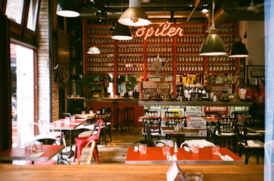 A cozy cafe interior with red accents, featuring a bar stocked with numerous bottles and vintage decor. Tables are set with neatly arranged glassware, and a warm, inviting atmosphere is enhanced by overhead lighting.