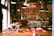 A cozy cafe interior with red accents, featuring a bar stocked with numerous bottles and vintage decor. Tables are set with neatly arranged glassware, and a warm, inviting atmosphere is enhanced by overhead lighting.