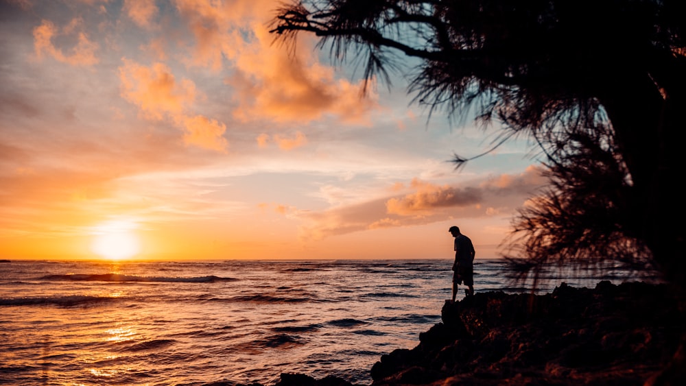 Silueta del hombre de pie sobre la roca cerca del cuerpo de agua durante la puesta del sol
